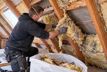 Isolatiemonteur verwijdert oude minerale wol tussen de houten dakbalken van een schuin dak